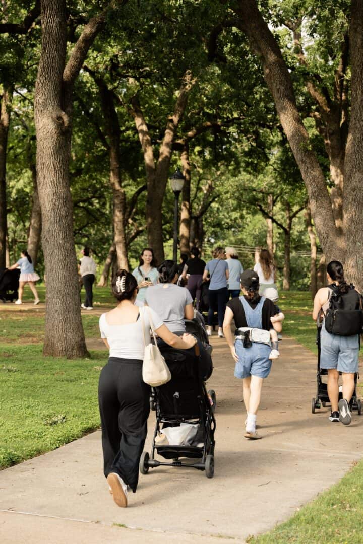 Moms walk along a paved path in a park surrounded by trees. Some moms push strollers, and children are visible. The scene takes place during daylight with greenery all around.