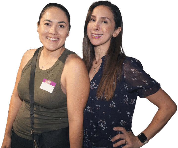 Two moms stand close together, smiling at the camera. One wears a sleeveless green top and crossbody bag with a name tag, while the other wears a short-sleeve floral blouse and an Apple Watch.