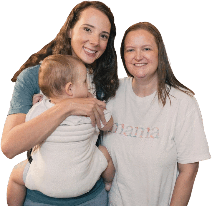 Two moms smile at the camera. One mom holds a baby who is facing away from the camera. The other mom is wearing a white shirt with the word "mama" printed on it.