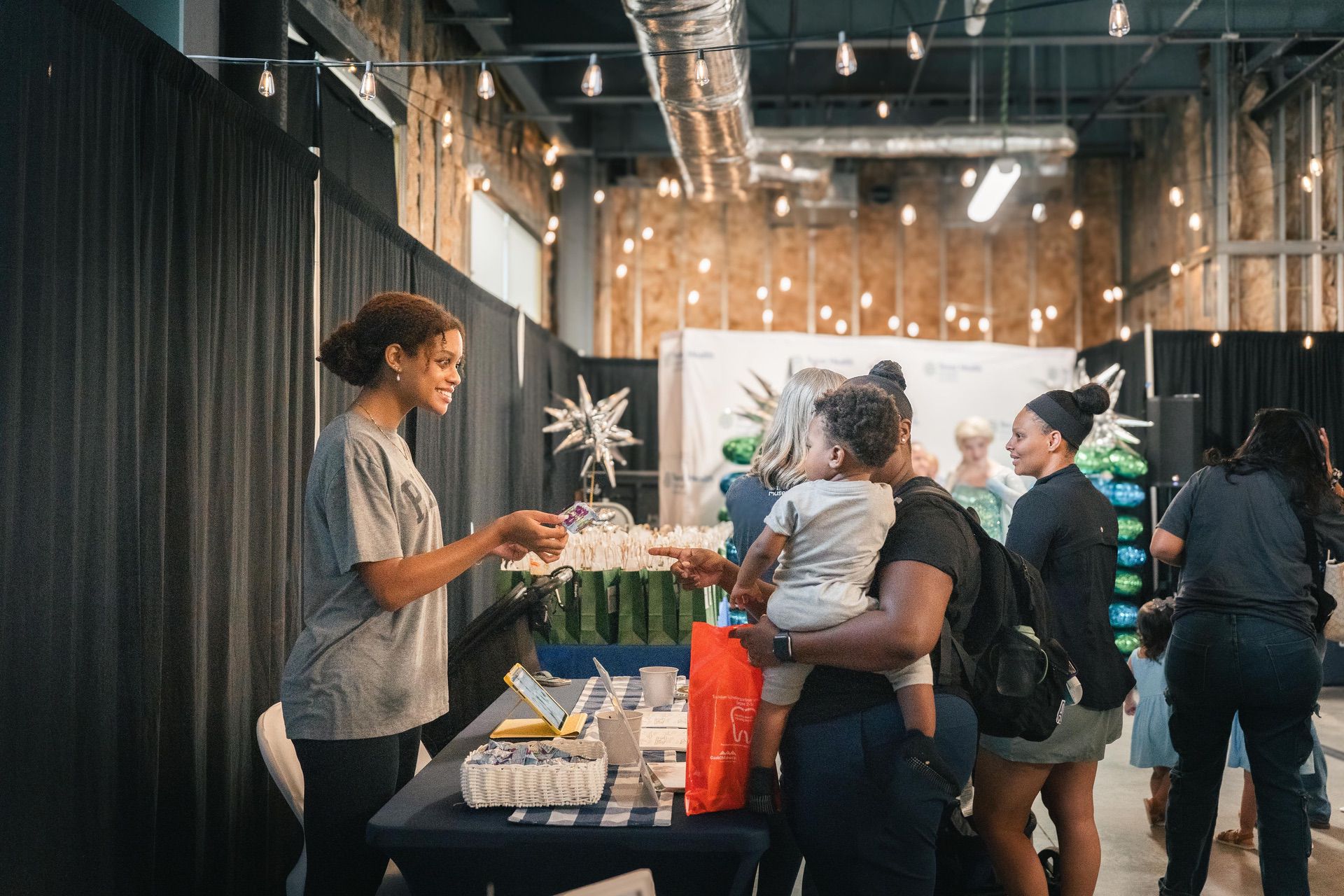 A woman stands behind a table at an indoor event, handing an item to another woman holding a child. Several people are gathered around, and balloons and decorations are visible in the background.