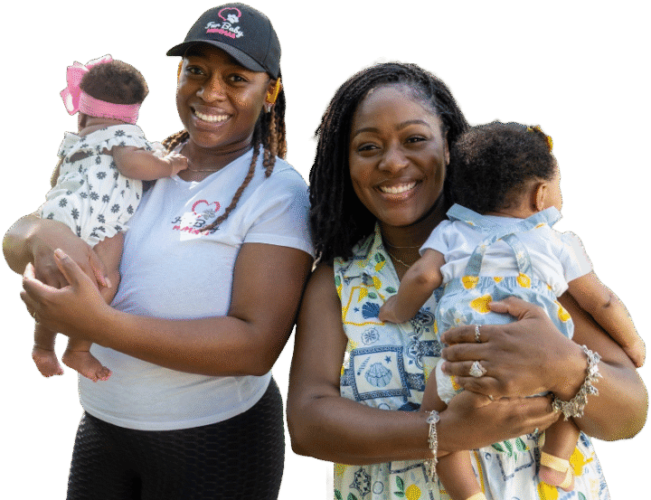 Two moms smile at the camera while each holding a baby. One mom wears a black cap and white shirt, and the other wears a patterned sleeveless top. Both babies have their backs to the camera.