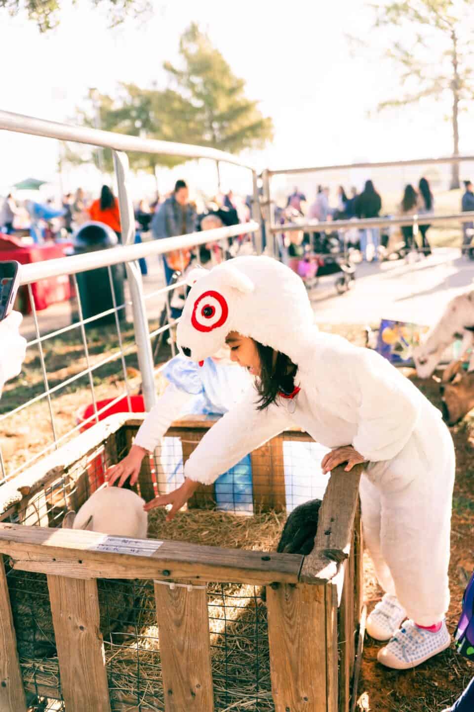 A child in a white animal costume reaches to pet a white rabbit inside a wooden enclosure at an outdoor event, with people and animals visible in the background.